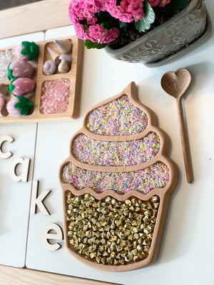 Cupcake-shaped wooden sensory tray filled with colorful rice and shiny gold beads with heart-shaped wooden spoon nearby