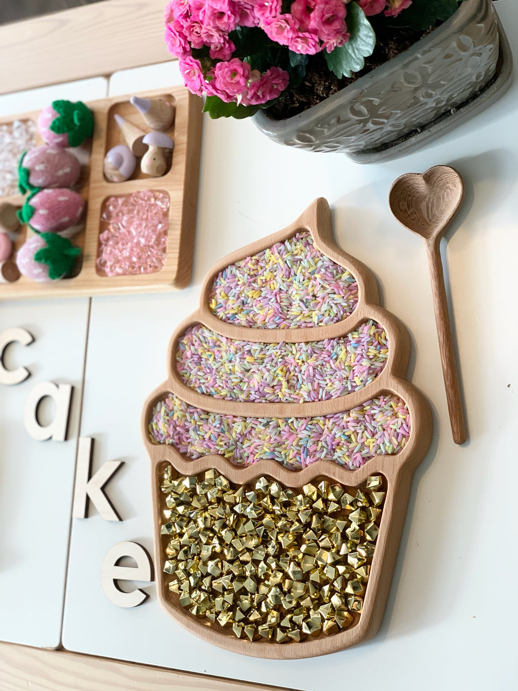 Cupcake-shaped wooden sensory tray filled with colorful rice and shiny gold beads with heart-shaped wooden spoon nearby