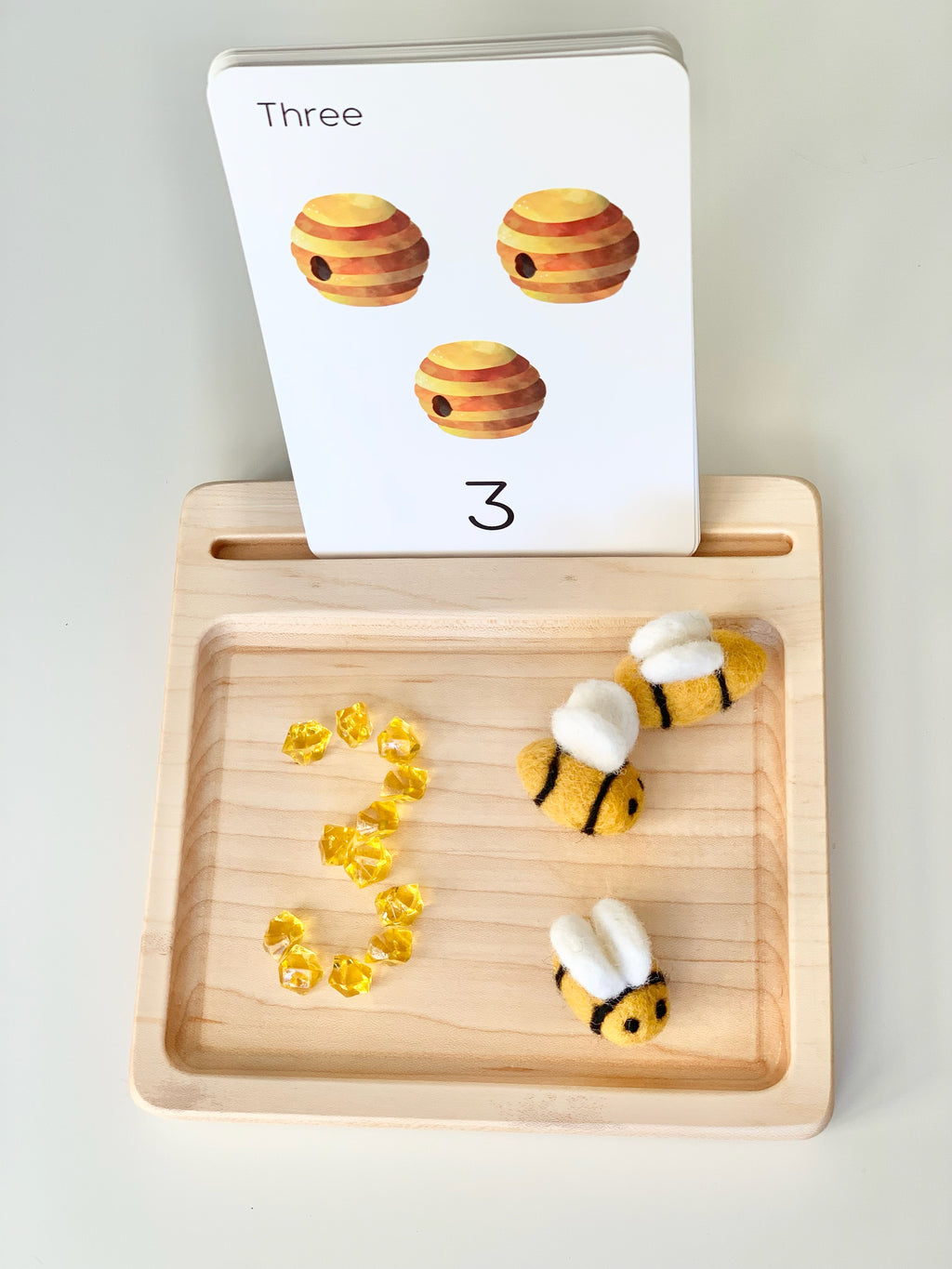 Wooden sensory tray with number 3 flash card, three bees, and yellow gems shaped in number three on light wood surface