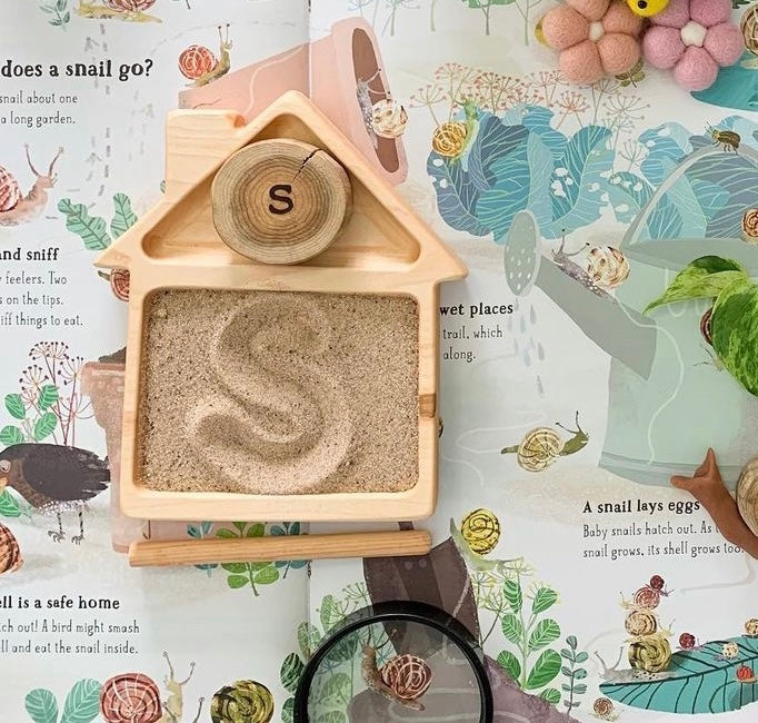 Natural wooden house-shaped sensory tray with sand and letter S stencil on illustrated nature background