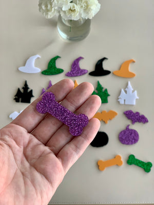 Hand holding purple glitter acrylic bone shape with various colorful Halloween acrylic shapes on white surface below