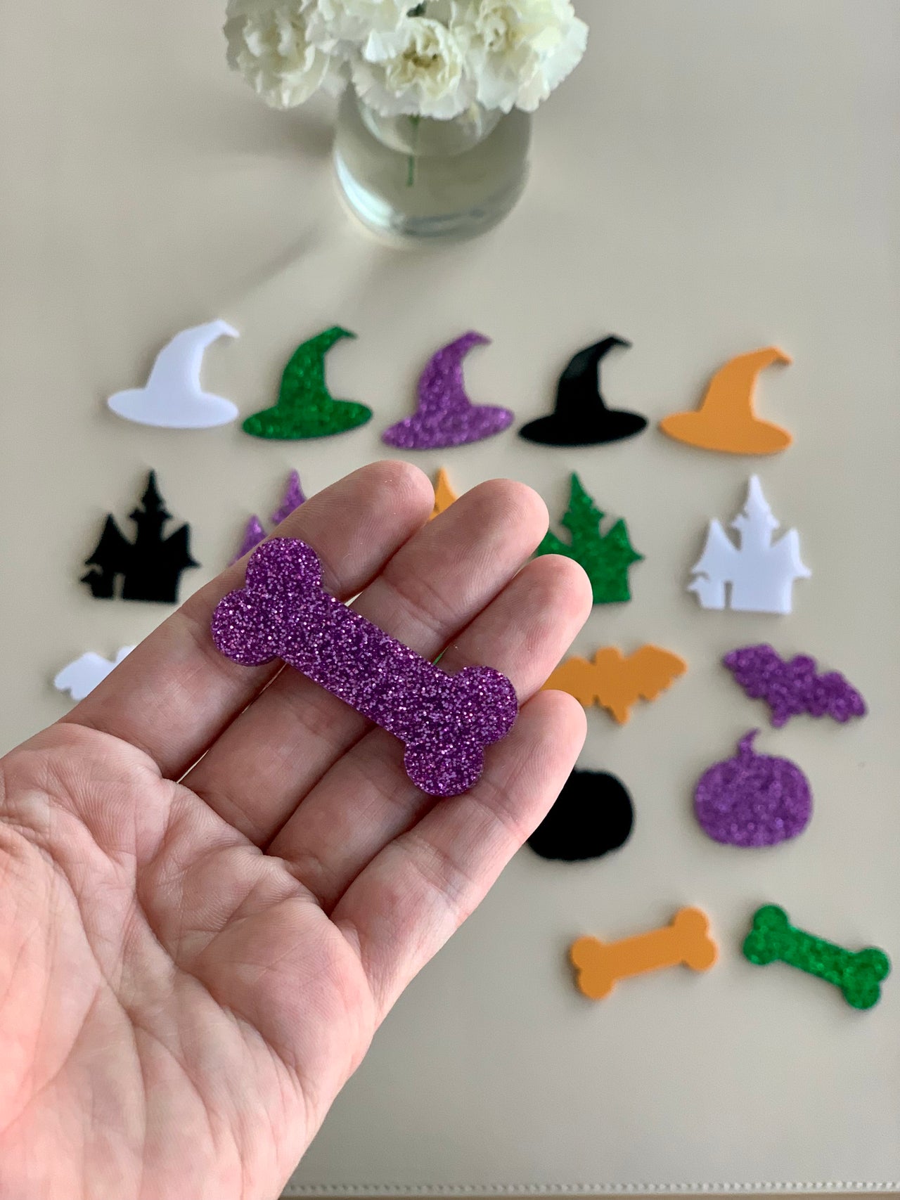 Hand holding purple glitter acrylic bone shape with various colorful Halloween acrylic shapes on white surface below