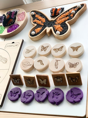 Wooden butterfly sensory tray filled with colored rice and set with wooden stamps and purple butterfly-shaped clay pieces on white surface