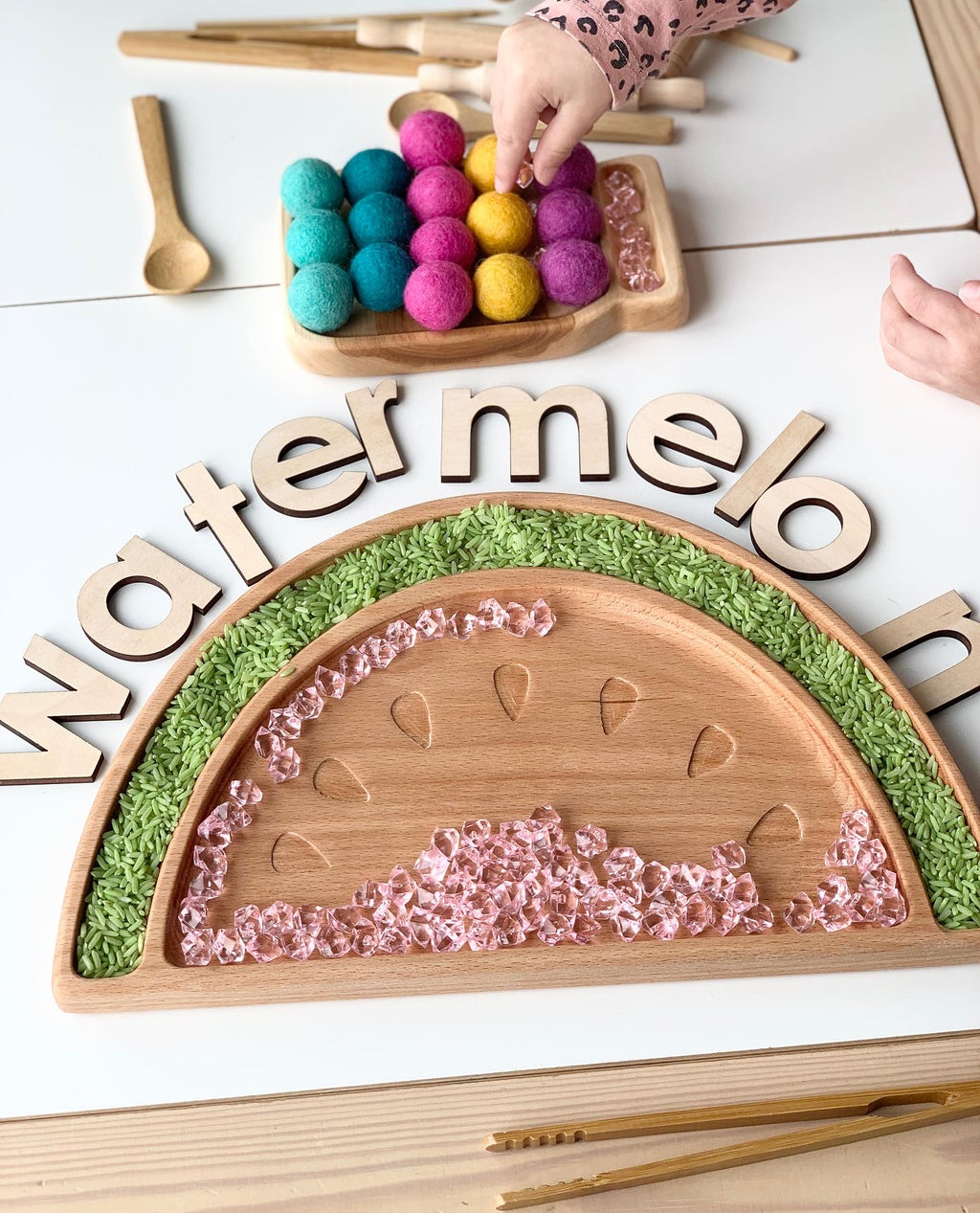Large wooden watermelon sensory tray with engraved seeds, green rice border, and clear pink beads on light wood table