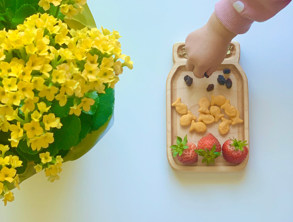 Small wooden jar-shaped sensory tray with snacks arranged inside and a child’s hand reaching for them on white surface