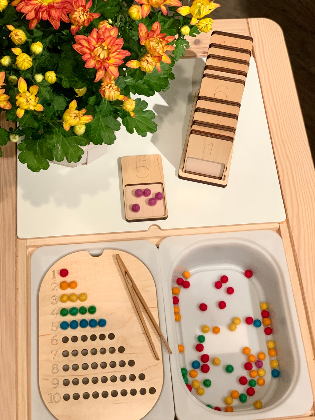 Wooden counting tray with colorful felt balls, number cards stacked on wooden stand, and tweezers on wooden table next to flowers