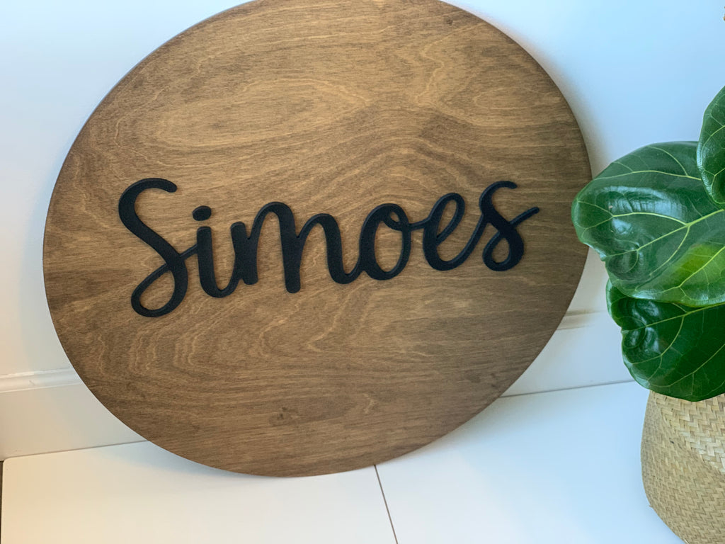 Round wooden 3D name sign with black cursive letters on natural wood background near green leafy plant on white floor