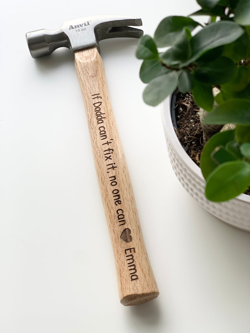 Personalized wooden handle hammer with laser engraved message and heart symbol beside green potted plant on white surface