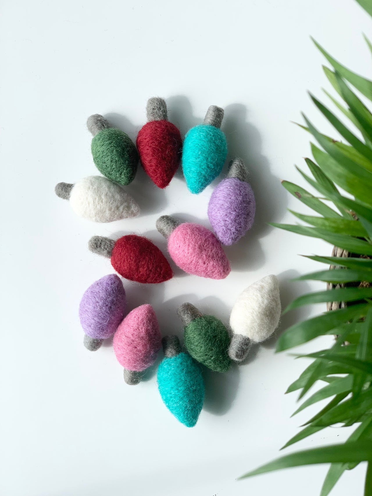 Colorful handmade felt Christmas light bulbs arranged in a circle on white background with green plant edge