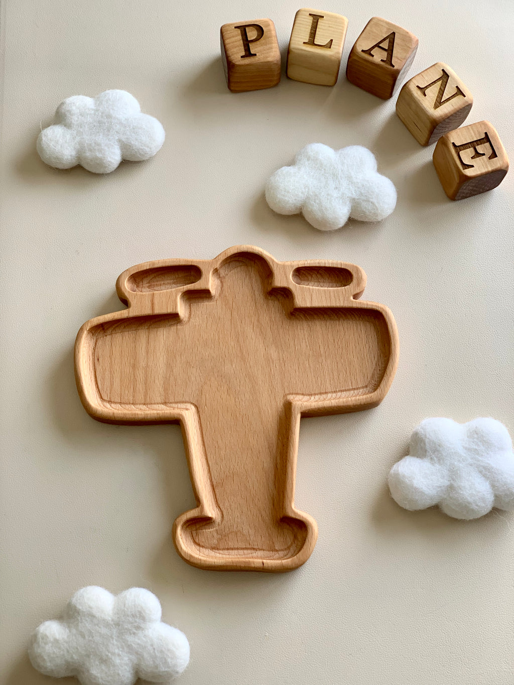 Wooden airplane-shaped sensory tray surrounded by felt clouds and wooden letter blocks spelling plane on beige surface