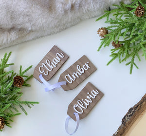 Three custom wooden 2x4 inch gift tags with white cursive names and satin ribbons on white surface with greenery decor