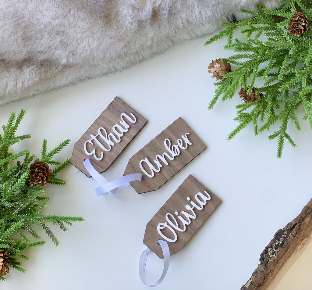 Three custom wooden 2x4 inch gift tags with white cursive names and satin ribbons on white surface with greenery decor