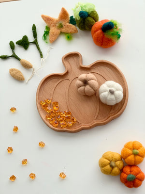 Natural wooden pumpkin-shaped plate with felt pumpkins, felt greenery, and amber-colored sensory beads on white background