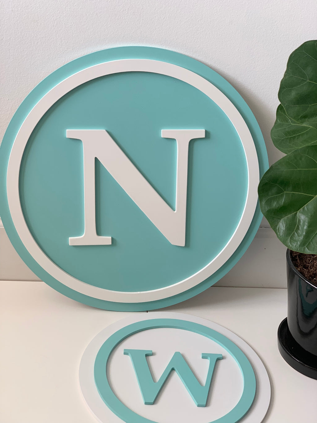 Round 3D monogram wooden sign with white letter N on turquoise background and decorative border next to small plant