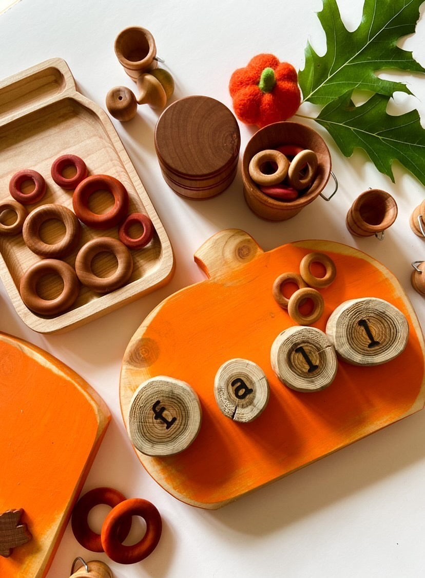 Small wooden sensory tray shaped like a pumpkin with engraved wood slices spelling "fall" and wooden rings on white background