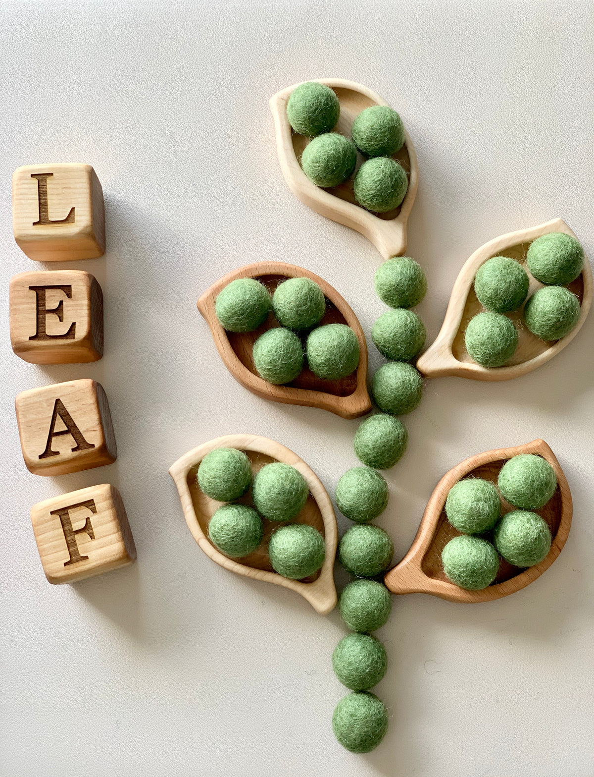 Small wooden leaf-shaped sensory trays filled with green felt balls arranged with wooden letter blocks spelling leaf on white surface