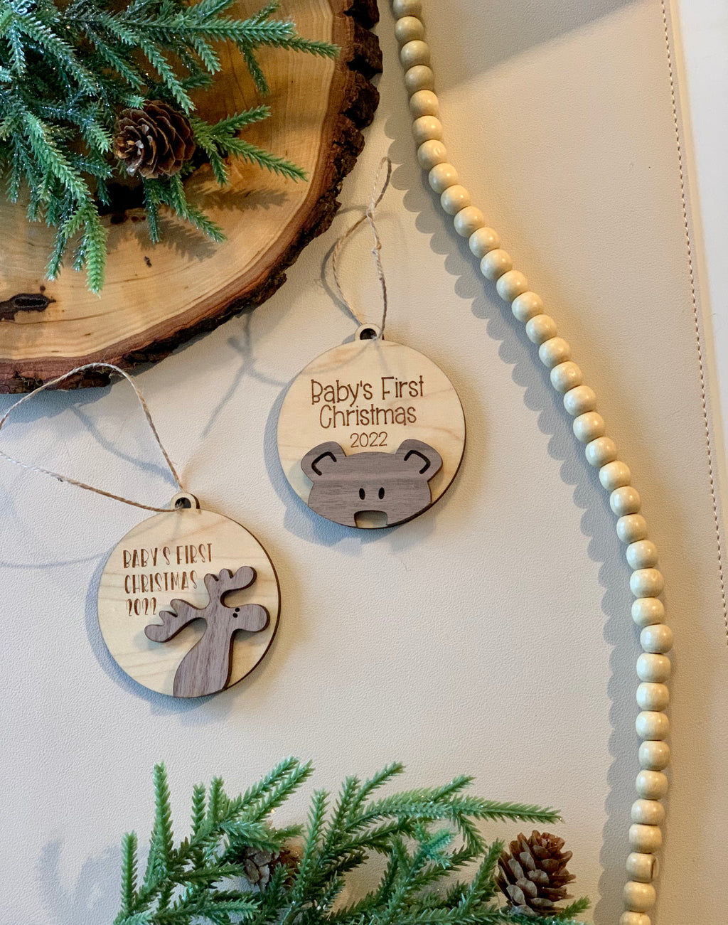Two wooden Baby’s First Christmas 2021 and 2022 ornaments with moose and bear designs on white surface