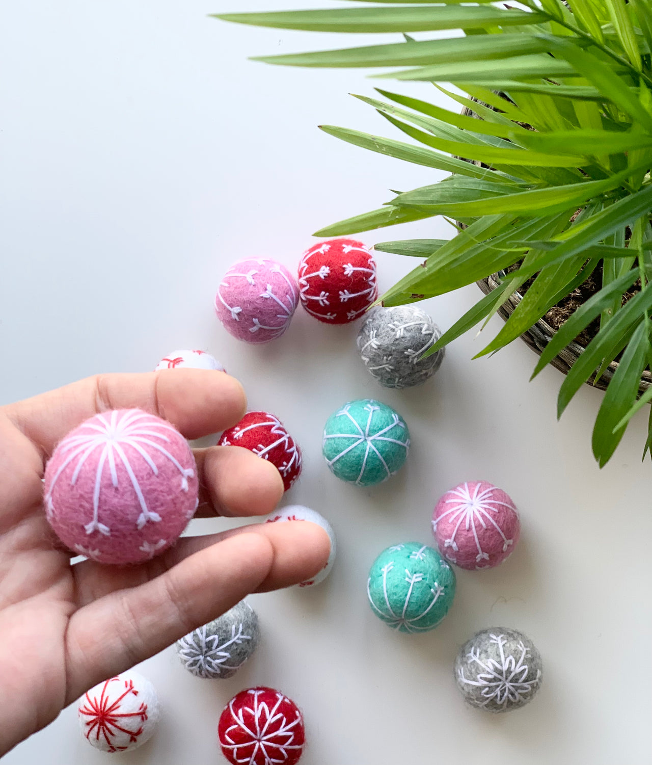 Hand holding a pink felt snowflake ball with white embroidery above other colorful felt snowflake balls on white surface near green plant