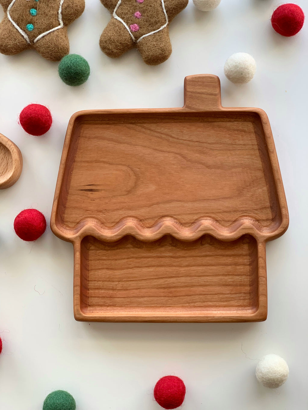 Wooden gingerbread house shaped plate with smooth finish surrounded by felt balls and gingerbread decorations on white surface