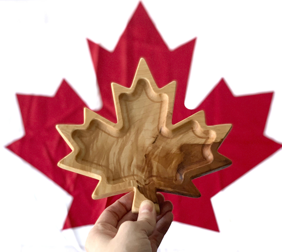 Hand-carved maple leaf wooden plate held in hand with red maple leaf flag background on white surface
