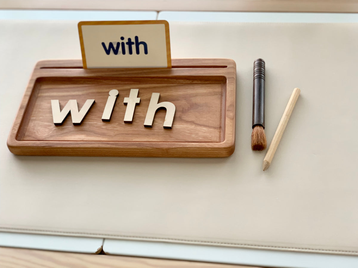 Rectangle wooden writing tray with wooden letters spelling "with," paintbrush and pencil on light background