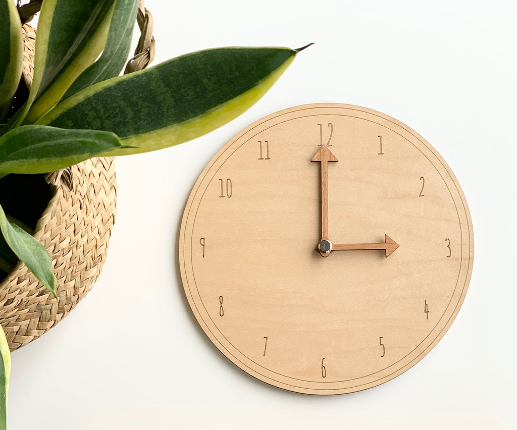 Round wooden play clock with movable hands and engraved numbers displayed beside green plant on white surface