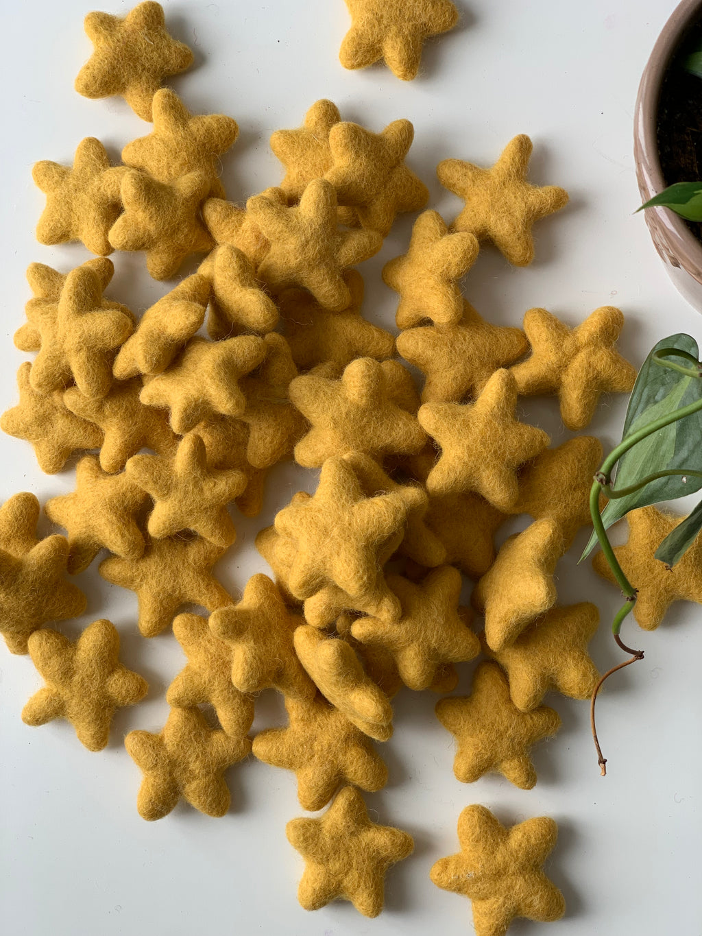 Multiple handmade mustard yellow felt stars scattered on white surface near a green potted plant
