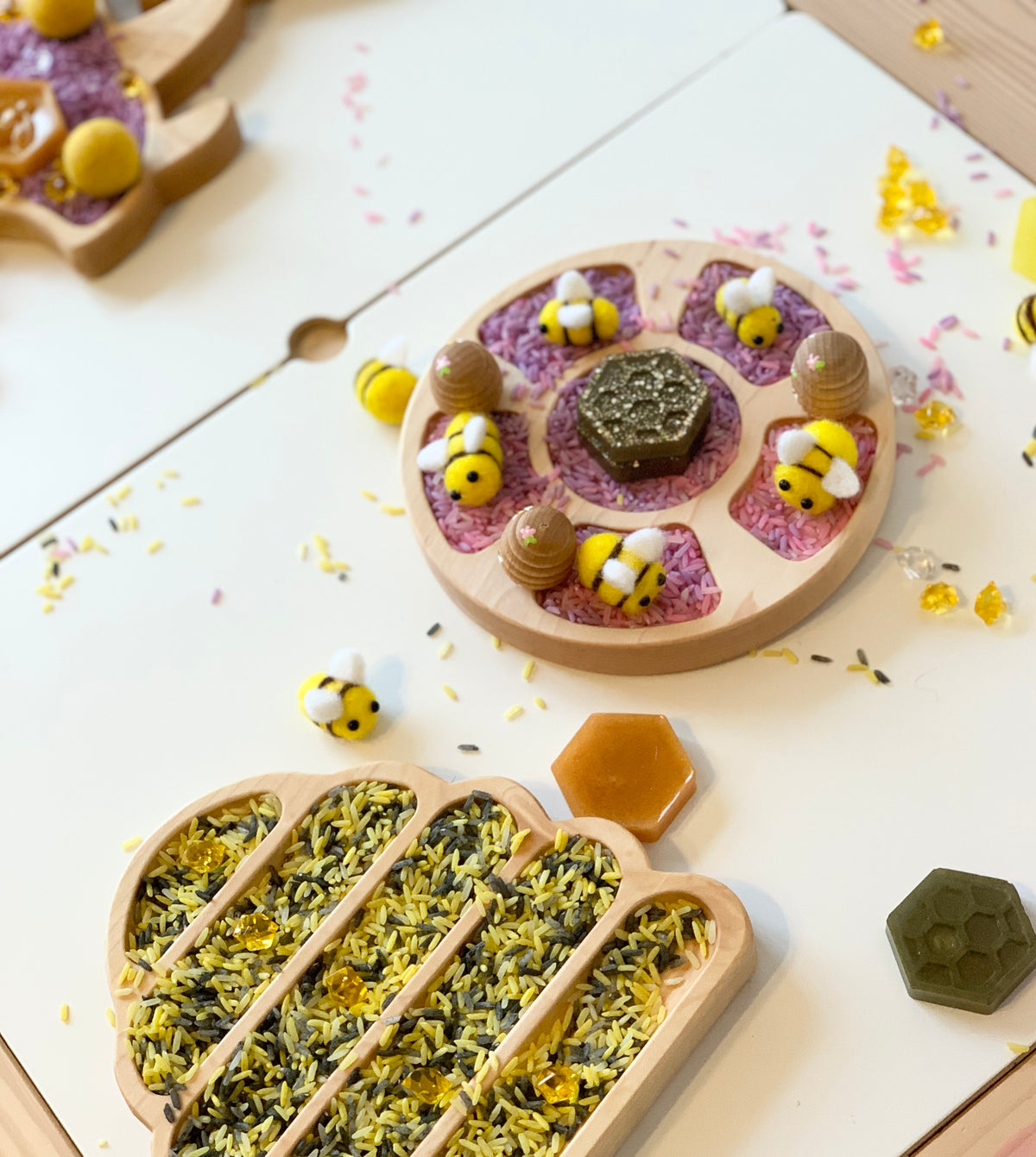 Wooden bee hive sensory trays filled with colorful rice and small bee and honeycomb shaped toys on white surface