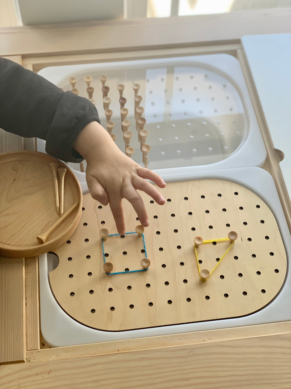Child hand placing rubber bands on pegs of small hole geometric light board tabletop insert on wooden table