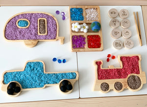 Wooden sensory trays shaped like a camper, truck, and train filled with colorful textured sensory materials on white surface