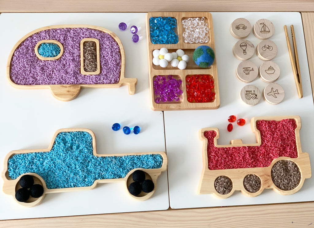 Wooden sensory trays shaped like a camper, truck, and train filled with colorful textured sensory materials on white surface
