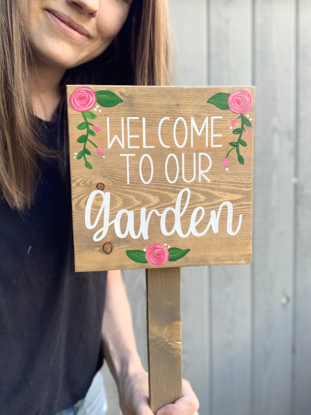 Small square wooden garden sign with white cursive lettering and pink floral accents held by person outdoors