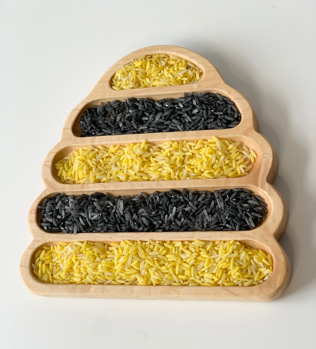 Natural wooden bee hive shaped sensory tray filled with alternating black and yellow rice on white background