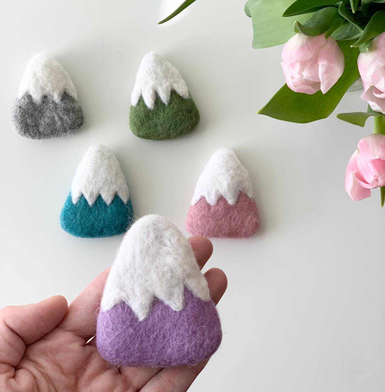 Handmade fair trade felt mountain in purple with white snowy peak held in hand with other colorful felt mountains nearby on white surface