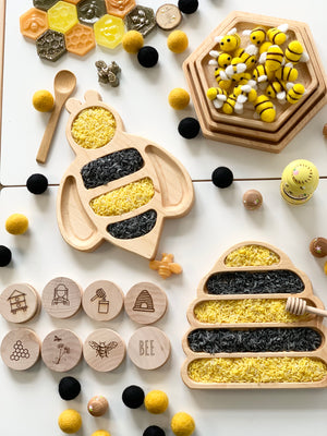 Natural wooden nesting hexagon plates and bee-shaped sensory trays filled with yellow and black rice on white background