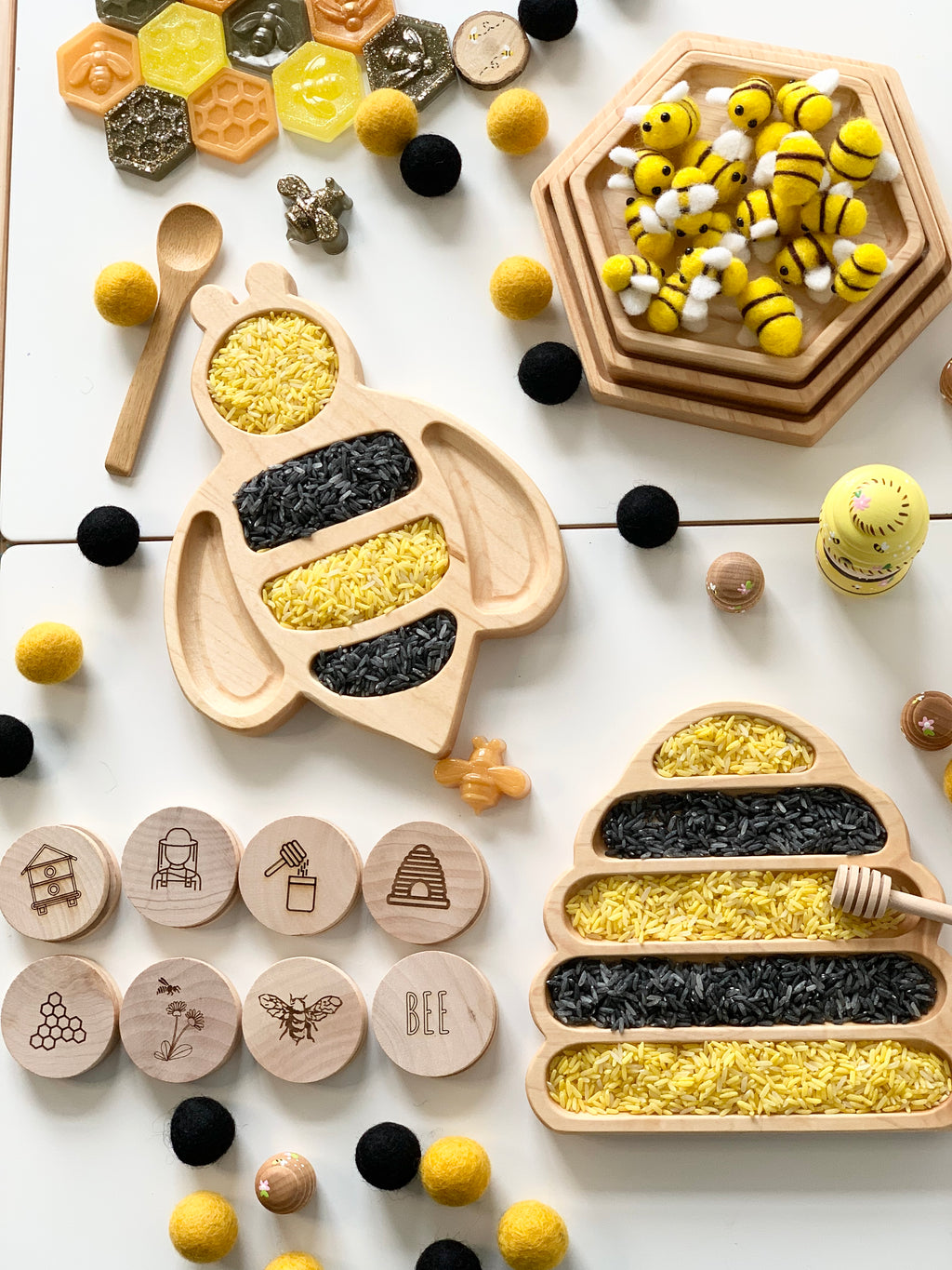 Natural wooden nesting hexagon plates and bee-shaped sensory trays filled with yellow and black rice on white background