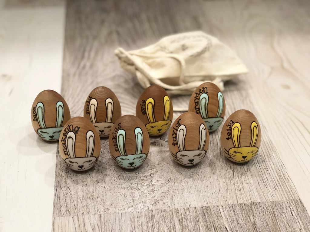Set of personalized wooden Easter eggs with painted bunny faces and names on natural wood background with cloth bag