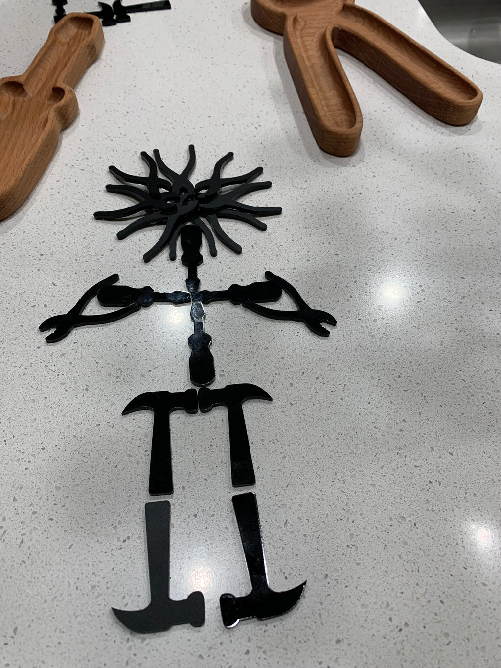 Black acrylic tool-shaped loose parts arranged in a figure shape on a white speckled surface with wooden trays nearby