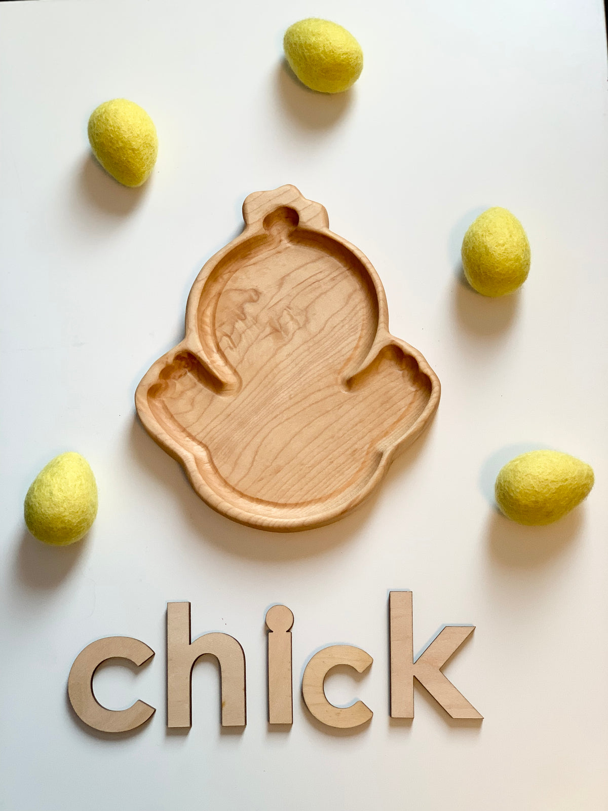 Wooden chick-shaped sensory tray surrounded by yellow felt eggs and wooden letters spelling chick on white surface