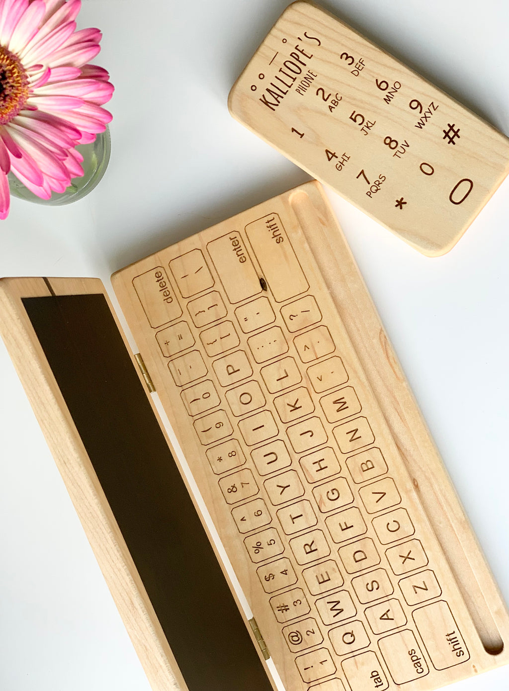 Wooden toy phone and laptop set with engraved numbers and letters on light natural wood surface with pink flower nearby