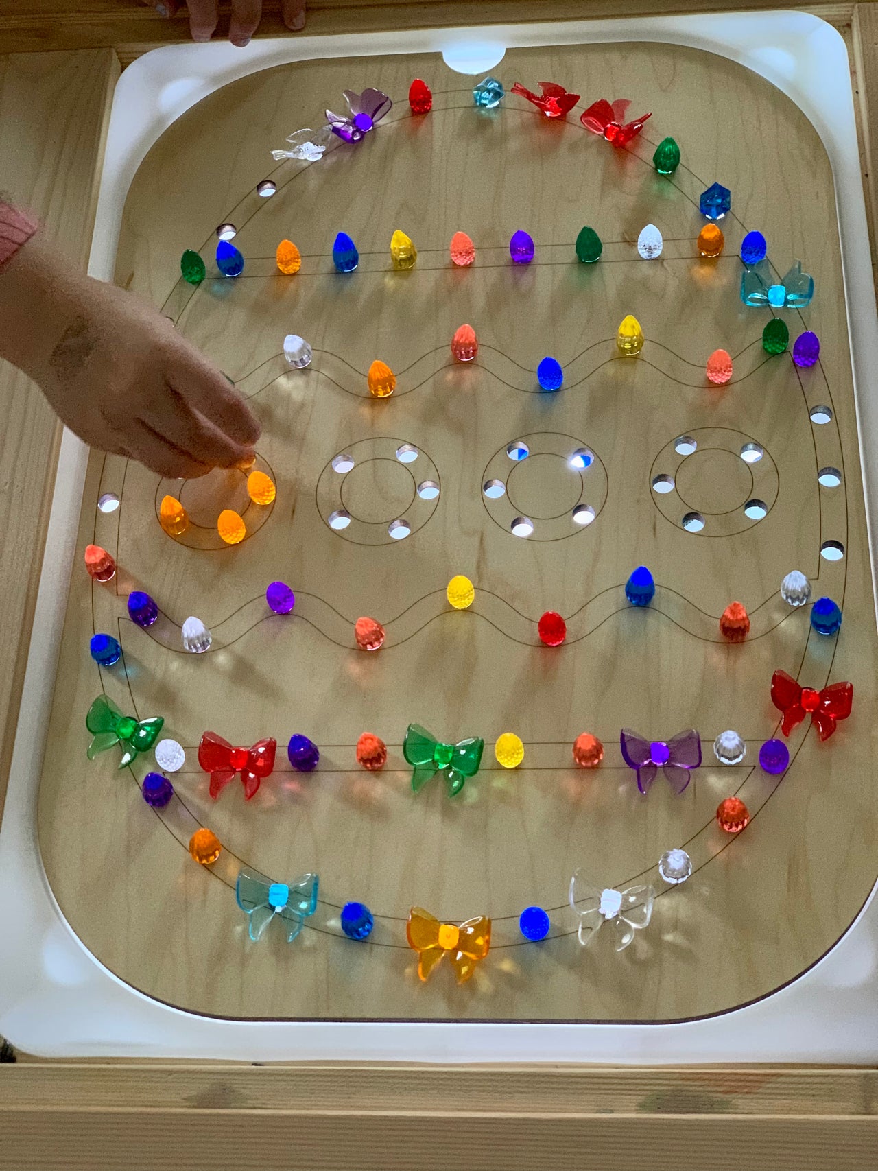 Child’s hand placing colorful light-up pegs shaped like eggs and butterflies into a wooden geometric light board on table