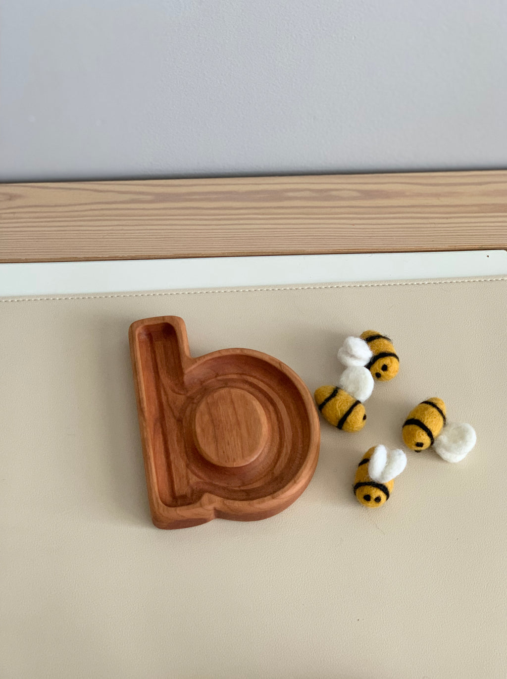 Natural wooden alphabet plate carved in the shape of letter b with small felt bees on beige surface