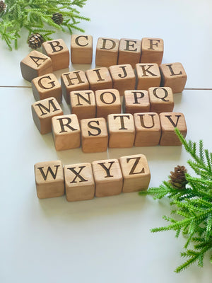 Natural 1.5-inch wooden alphabet blocks with engraved capital letters arranged on white surface with green foliage and pinecones