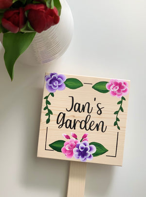 Small natural wooden square garden sign with Jan's garden in black cursive and hand-painted pink and purple flowers with green leaves