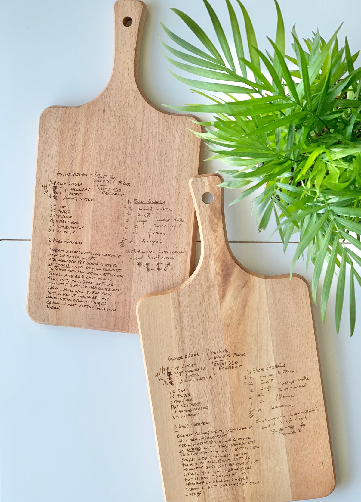 Two personalized wooden cutting boards with laser engraved handwritten recipes on light wood beside green plant