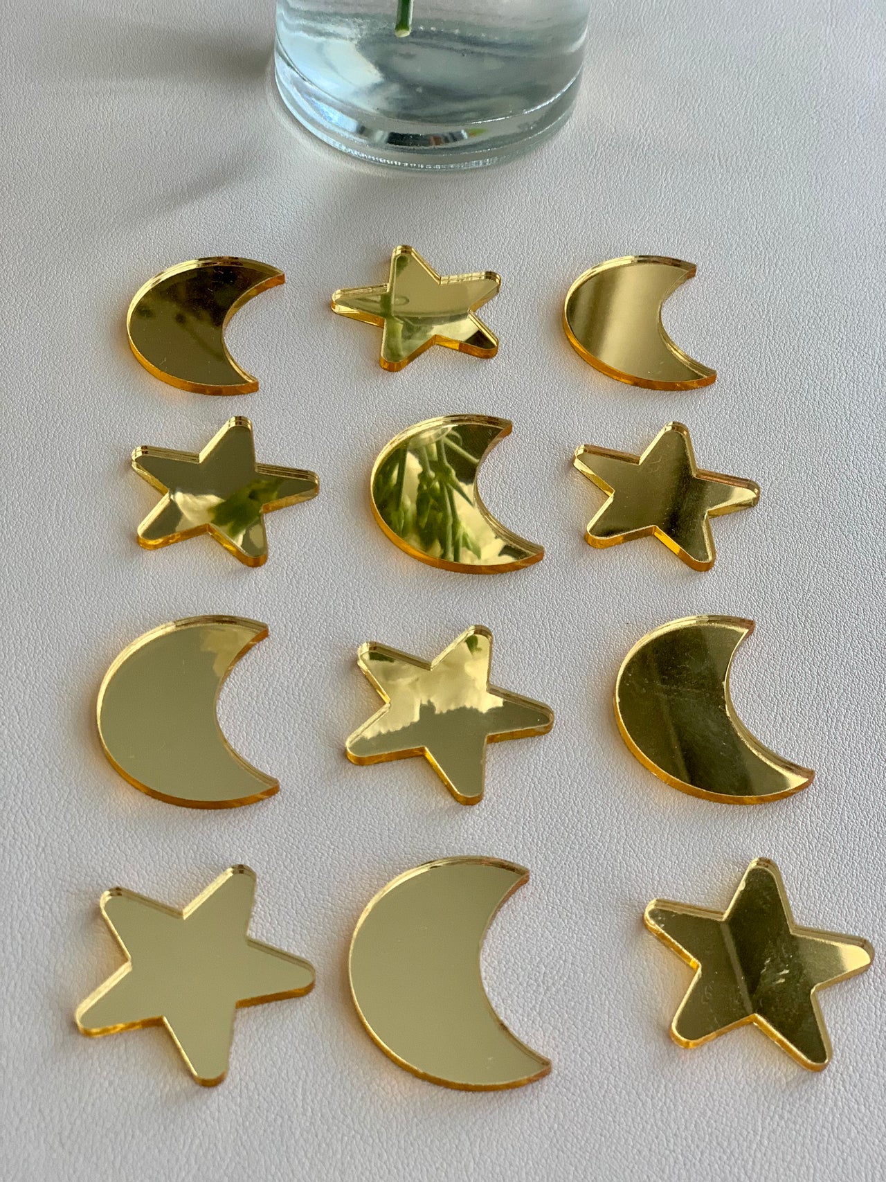Set of 12 gold acrylic loose parts shaped like moons and stars arranged on white surface below glass vase