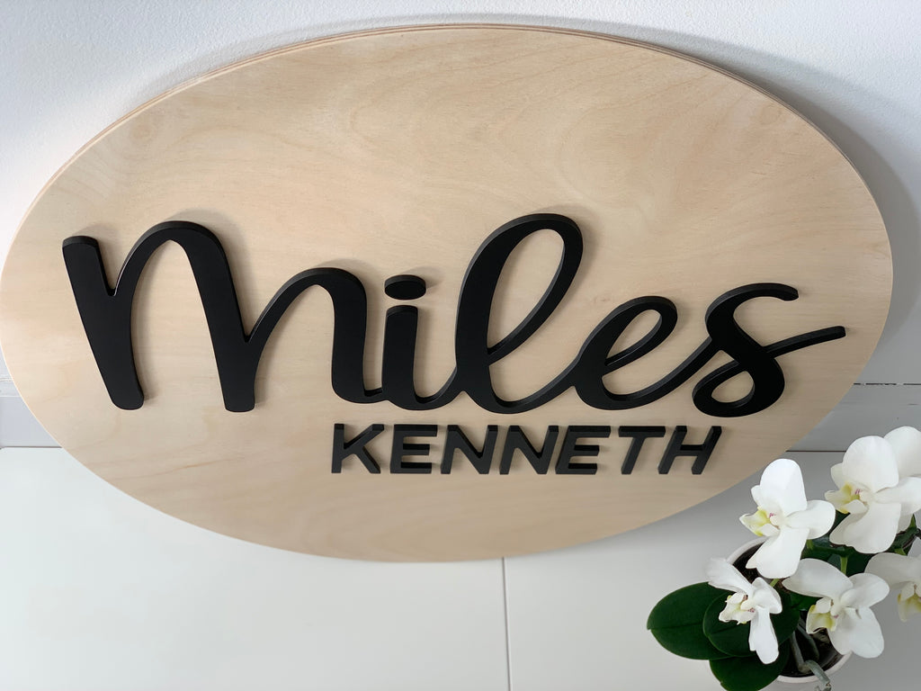 Oval natural wooden 3D name sign with black cursive "Miles" and block "Kenneth" letters on white surface with white orchid plant