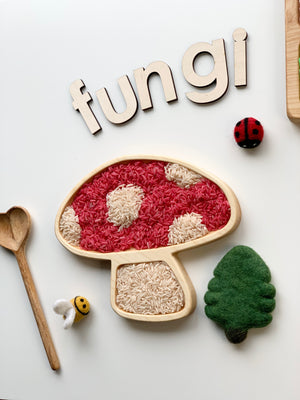Wooden tray shaped like a mushroom filled with red and white rice next to felt ladybug, felt tree, wooden spoon, and wooden letters spelling fungi