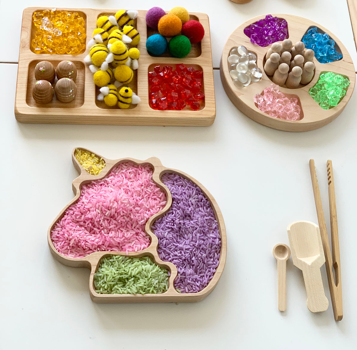 Natural wooden unicorn sensory tray filled with colorful dyed rice next to wooden sensory play tools and trays with beads and felt balls