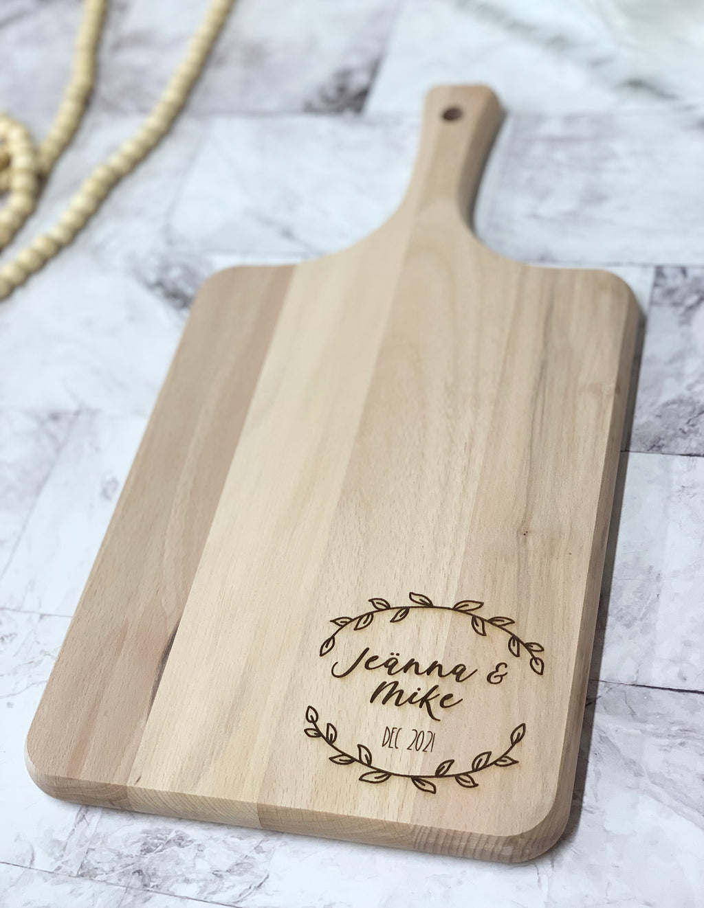 Natural wooden personalized cutting board with engraved names and decorative leaf wreath on light wood surface